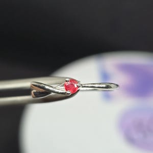 Puede incluir: Un delicado anillo de plata con una pequeña gema redonda de color rojo brillante. La banda del anillo tiene un diseño retorcido, añadiendo un toque de elegancia. La gema está engastada de forma segura, mostrando su color vibrante. El anillo es sujetado por unas pinzas.