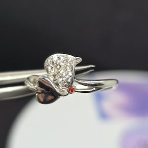 May include: A silver ring featuring a floral design. The ring's focal point is a cluster of small, clear gemstones, with a single red gemstone accent. The ring is held by a pair of silver tweezers against a blurred background.