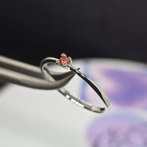 May include: A delicate silver ring featuring a small, orange gemstone cluster. The ring is held by a pair of silver tweezers, showcasing its intricate design. The gemstone is a vibrant orange color, set against the polished silver band.