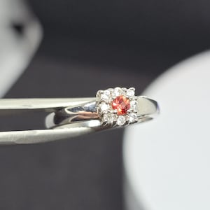 Könnte beinhalten: Ein silberner Ring mit einem zentralen orangefarbenen Edelstein, umgeben von kleineren, klaren Steinen. Der Ring wird von einer Pinzette vor einem verschwommenen Hintergrund gehalten. Das Band des Rings ist glatt und poliert.