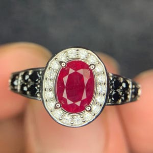 Peut inclure: Une bague avec une pierre rouge centrale de taille ovale entourée d'un halo de petites pierres blanches. L'anneau est en métal foncé et orné de grappes de petites pierres noires de chaque côté du sertissage central.