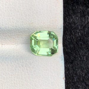 May include: A light green, emerald-cut gemstone with a rectangular shape and faceted edges. The gem is set against a white surface, with a dark, textured background. The gemstone has a bright, translucent appearance.