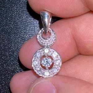 May include: A silver pendant with a double-circle design. The top circle is smaller and has a textured surface. The bottom circle is larger and features a central stone surrounded by smaller stones. The pendant is held by a person's fingers.