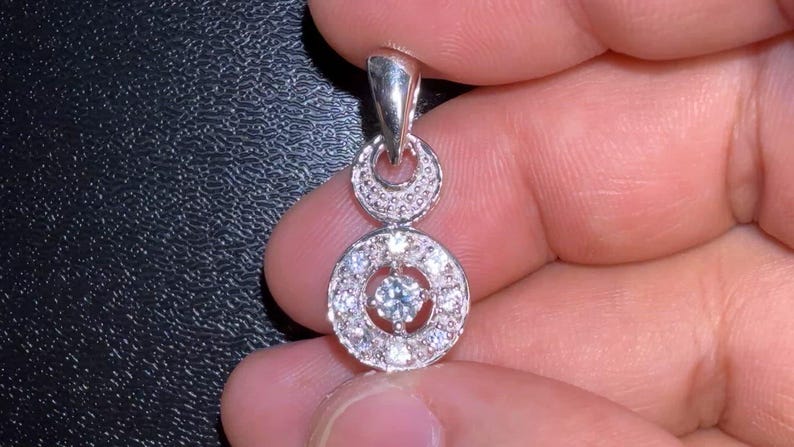 May include: A silver pendant with a double-circle design. The top circle is smaller and has a textured surface. The bottom circle is larger and features a central stone surrounded by smaller stones. The pendant is held by a person's fingers.
