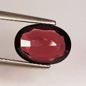 以下が含まれることがあります： 銀色のピンセットで保持された、ファセット加工された楕円形の濃い赤色の宝石。 宝石は、光を反射して輝く効果を生み出す複数のファセットを持つ研磨された表面を持っています。 宝石の色は、豊かで濃い赤色です。
