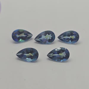 May include: Five pear-shaped, faceted gemstones with a blue-green color. The gems are arranged on a white surface. Each gem has internal reflections, creating a play of light and color. The gems are likely for jewelry making or collecting.