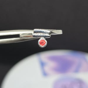 Puede incluir: Un anillo de plata con una pequeña gema redonda de color naranja. El anillo tiene un diseño minimalista con un detalle rectangular y pequeños adornos. El anillo es sujetado por unas pinzas contra un fondo borroso.