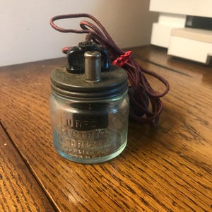 Könnte beinhalten: Ein antiker Glasbehälter mit schwarzem Deckel und einer Metalldüse. Der Behälter ist mit "Turco Electric Vaporizer" beschriftet und hat ein rot-schwarzes Stromkabel.
