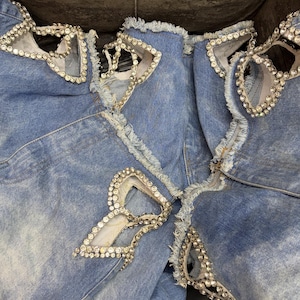 May include: Light blue denim jeans featuring multiple distressed cutouts with frayed edges. Each cutout is intricately bordered with clear, sparkling rhinestones that follow the shape of the openings, creating a decorative and textured appearance.