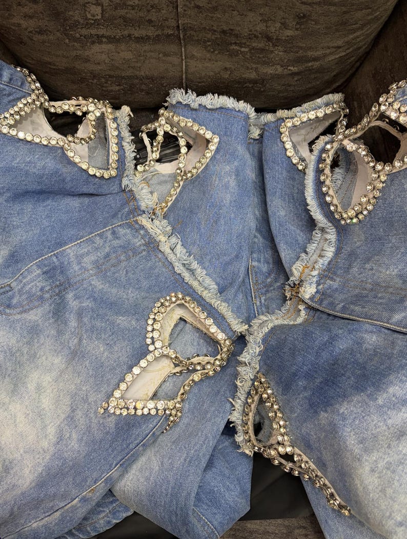 May include: Light blue denim jeans featuring multiple distressed cutouts with frayed edges. Each cutout is intricately bordered with clear, sparkling rhinestones that follow the shape of the openings, creating a decorative and textured appearance.
