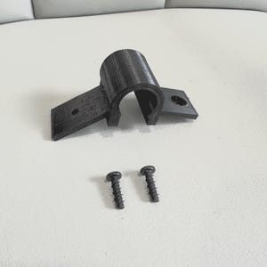 May include: A black 3D-printed pipe clamp with two screws. The clamp has a curved section and two flat extensions with holes for mounting. The screws are dark and have a threaded design.