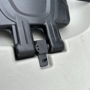 May include: Close-up of a black plastic seat latch on a white kayak. The latch is secured with a black strap.