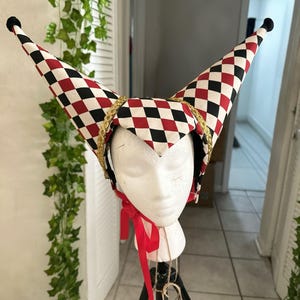 May include: A jester's hat with a red, black, and white checkered pattern. The hat has two tall, pointed tips with black pom-poms. Gold trim and a red ribbon add detail to the hat, displayed on a mannequin head.