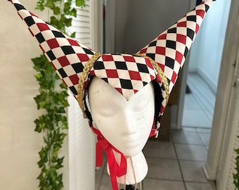 May include: A jester's hat with a red, black, and white checkered pattern. The hat has two tall, pointed tips with black pom-poms. Gold trim and a red ribbon add detail to the hat, displayed on a mannequin head.