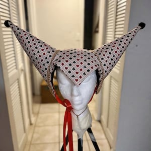 May include: A two-pointed jester-style hat featuring a white fabric base with a repeating pattern of red hearts, red diamonds, black clubs, and black spades. The hat includes gold-toned trim along the edges and a bright red ribbon chin strap.