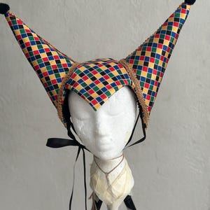 May include: A two-pointed jester hat featuring a colorful diamond pattern in red, yellow, blue, green, and black. The hat is trimmed with gold braided cord, features black pom-poms at each tip, and includes black ribbon ties for securing.