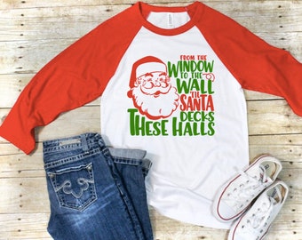May include: White and red baseball tee shirt with a green graphic of Santa Claus and the text "From the window to the wall til Santa decks these halls".