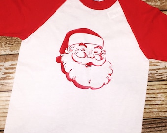 May include: White and red raglan baseball tee featuring a red Santa Claus face graphic. The Santa Claus graphic is centered on the white body of the shirt. The sleeves and neckline are red.