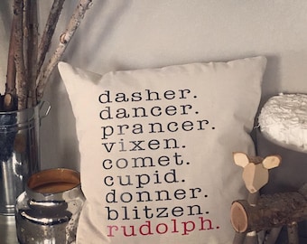 May include: A white pillow with black text listing the names of Santa's reindeer: Dasher, Dancer, Prancer, Vixen, Comet, Cupid, Donner, Blitzen, and Rudolph. The word Rudolph is in red.