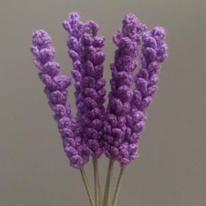 Op de afbeelding: Een boeket handgemaakte lavendelbloemen in paarse tinten. De gehaakte bloemen hebben gedetailleerde knoppen en worden bij elkaar gehouden door groene stelen. De stelen zijn omwikkeld met groen garen. De achtergrond is effen grijs.