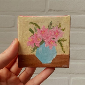 Op de afbeelding: Een klein vierkant schilderij van roze bloemen in een lichtblauwe vaas. De vaas staat op een bruin oppervlak tegen een beige achtergrond. Groene bladeren en stelen zijn zichtbaar. Het schilderij wordt door een hand vastgehouden.