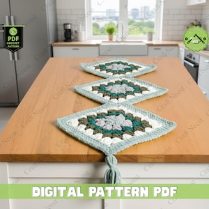 May include: Three square crochet placemats in green, white, and grey are displayed on a wooden kitchen island. Each placemat has a tassel. The text "DIGITAL PATTERN PDF" is at the bottom of the image.