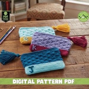 Pode incluir: Quatro bolsas de crochê feitas à mão em tons de azul, roxo e amarelo, cada uma com uma borla e uma letra. As bolsas estão dispostas sobre uma superfície de madeira, com agulhas de crochê e tesouras por perto. O texto "DIGITAL PATTERN PDF" está na parte inferior.