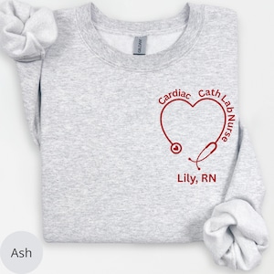May include: Ash grey sweatshirt with a red heart and stethoscope graphic. The text "Cardiac Cath Lab Nurse" is written around the heart and "Lily, RN" is written below the heart.