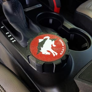 Peut inclure: Un couvercle de bouton noir circulaire avec un centre rouge texturé, une silhouette de cheval cabré blanche et le texte G.O.A.T. MODES imprimé en blanc. Le bouton présente un bord extérieur cranté et est situé sur la console centrale du véhicule.