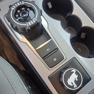 May include: A black gear shift knob with white text reading BRONCO SPORT and gear positions P, R, N, D, L. Below it, a black parking brake button and a steering wheel icon button. A secondary black knob features a white bucking horse logo.