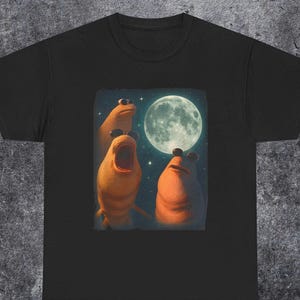 Pode incluir: Camiseta preta com uma ilustração fantasiosa de três criaturas laranja olhando para uma lua cheia em um céu estrelado. As criaturas têm olhos grandes e diferentes expressões.
