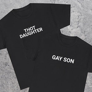 May include: Two black t-shirts with white text. One shirt reads "THOT DAUGHTER" and the other reads "GAY SON". The shirts are made of a soft material and have a classic crew neck design.
