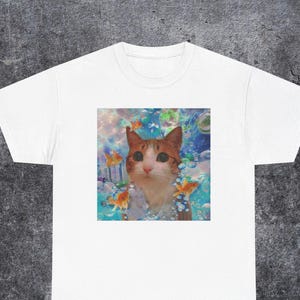 Puede incluir: Una camiseta blanca de manga corta con un estampado gráfico centrado de un gato atigrado naranja con grandes ojos oscuros, rodeado de peces dorados y burbujas azules en un entorno submarino con una paleta de colores suave y etérea.