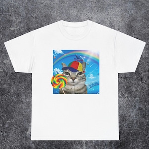 Puede incluir: Camiseta blanca con un gráfico caprichoso de un gato con una gorra colorida y sosteniendo una piruleta de arcoíris. El gráfico incluye un arcoíris, delfines y un fondo de cielo azul.