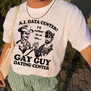 May include: A white short-sleeved t-shirt featuring a black graphic print of two men shaking hands. The text reads: A.I. DATA CENTER? I'd rather be at the... GAY GUY DATING CENTER. The shirt has a crew neckline and a standard fit.