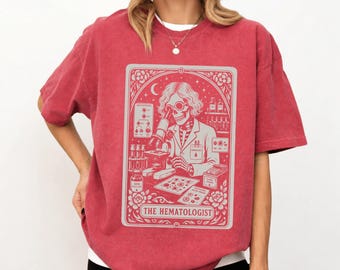 May include: A red, acid-washed t-shirt featuring a light gray graphic print of a skeleton scientist looking through a microscope. The design includes laboratory equipment, test tubes, and the text THE HEMATOLOGIST framed by floral borders.