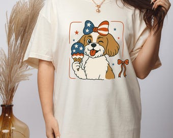 May include: A white t-shirt featuring a cartoon image of a dog wearing a red, white and blue bow and holding an ice cream cone. The dog is winking and has its tongue sticking out. The image is surrounded by a red frame and stars.