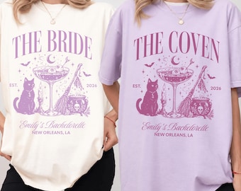 May include: Two oversized t-shirts, one cream and one light purple, featuring a purple graphic print. The design includes a cat, a cocktail glass with eyeballs, a broom, a cauldron with a skull, and text reading THE BRIDE or THE COVEN, EST. 2026, Emily's Bachelorette, NEW ORLEANS, LA.