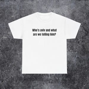 May include: A white short-sleeved t-shirt featuring the black printed text Who's aviv and what are we telling him? centred on the chest. The garment has a classic crew neckline and a standard fit, providing a clean and simple appearance.