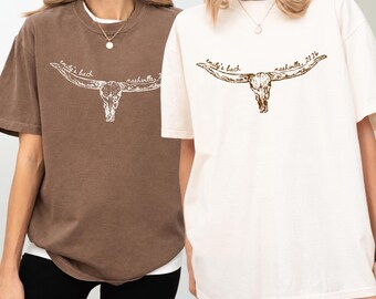 May include: A brown t-shirt and a cream t-shirt, each featuring a graphic of a longhorn skull with the text "emily's bach" and "nashville, 2026" printed in a cursive script above the horns. Both shirts have a crew neckline and short sleeves.
