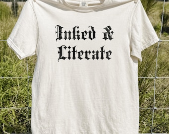 May include: An ivory short-sleeve t-shirt featuring the black text Inked & Literate in a gothic-style font centered on the chest. The shirt has a crew neckline and is displayed hanging from a clothesline against a natural outdoor setting.
