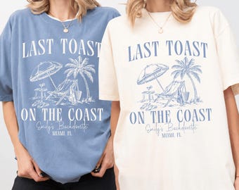 May include: Two short-sleeve t-shirts, one blue and one cream, each featuring a graphic print of a beach scene with a lounge chair, umbrella, and palm tree. The text reads LAST TOAST ON THE COAST, Emily's Bachelorette, and MIAMI, FL.