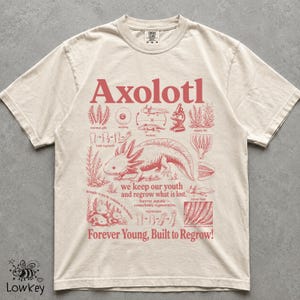 May include: A cream-coloured short-sleeve t-shirt featuring a red graphic print of an axolotl surrounded by scientific diagrams and text including "Axolotl," "we keep our youth and regrow what is lost," and "Forever Young, Built to Regrow!"