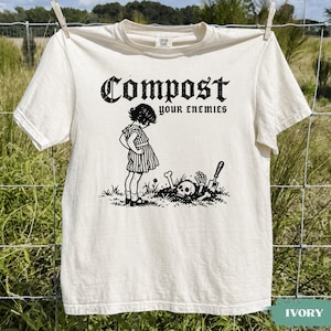 May include: An ivory short-sleeve t-shirt featuring a black graphic print of a child standing over a skull and bones in the dirt. The text above the illustration reads "Compost your enemies" in a bold, distressed black gothic-style font.