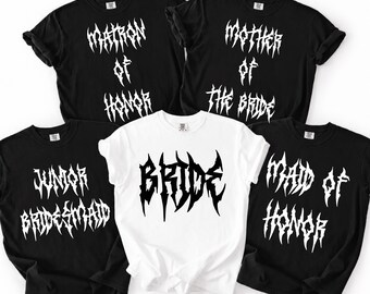 May include: A collection of five short-sleeved t-shirts featuring heavy metal style lettering. Four black shirts display white text reading Matron of Honor, Mother of the Bride, Junior Bridesmaid, and Maid of Honor. One white shirt reads Bride.