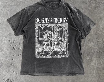 May include: A charcoal gray short-sleeve t-shirt with a faded wash. The front features a white graphic of two knights in armor holding swords and hands, framed by a forest border. Above the illustration, the text reads BE GAY & MERRY.