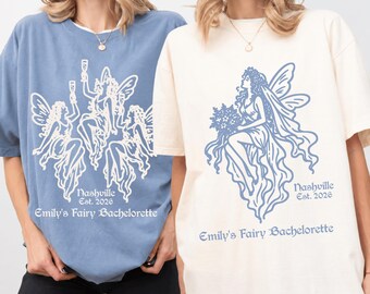 May include: A blue t-shirt with a white graphic of three fairies holding glasses and a cream t-shirt with a blue graphic of a single fairy holding flowers. Both shirts feature the text Nashville Est. 2026 and Emily's Fairy Bachelorette.