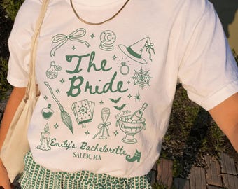 May include: A white t-shirt featuring dark green line art graphics including a witch hat, crystal ball, broom, spider web, and champagne bucket. The text reads The Bride, Emily's Bachelorette, EST. 2026, Champagne, and SALEM, MA.