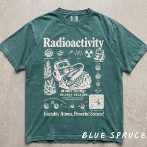 Peut inclure: Un t-shirt bleu-vert à manches courtes avec une impression graphique couleur crème sur la radioactivité. Le motif comprend des illustrations scientifiques d'atomes, de cristaux, d'un compteur Geiger et le texte Radioactivity, atoms change energy escapes, et Unstable Atoms, Powerful Science!