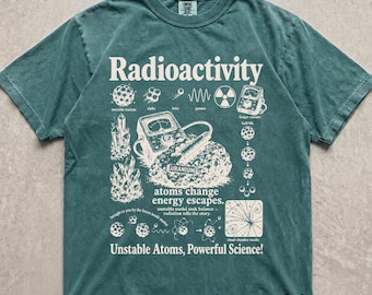 May include: A blue-green short-sleeve t-shirt featuring a cream-colored graphic print about radioactivity. The design includes scientific illustrations of atoms, crystals, a Geiger counter, and text reading Radioactivity, atoms change energy escapes, and Unstable Atoms, Powerful Science!